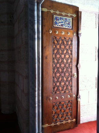 A beautifully carved wooden door with intricate geometric patterns. The top of the door features an ornate inscription in Arabic script. The door is framed by a stone wall, allowing a glimpse of an interior space with a red carpeted floor.