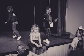 A group of children are on a stage, either standing or playing. One child in the center appears to be holding a certificate or piece of paper while others are moving around. The mood seems casual and playful, typical of a children's performance or play.