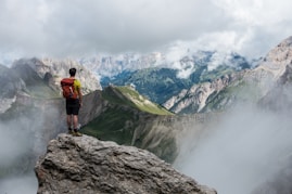 man with red backpack standing on cliff facing mountains under white sky during daytime