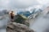man with red backpack standing on cliff facing mountains under white sky during daytime