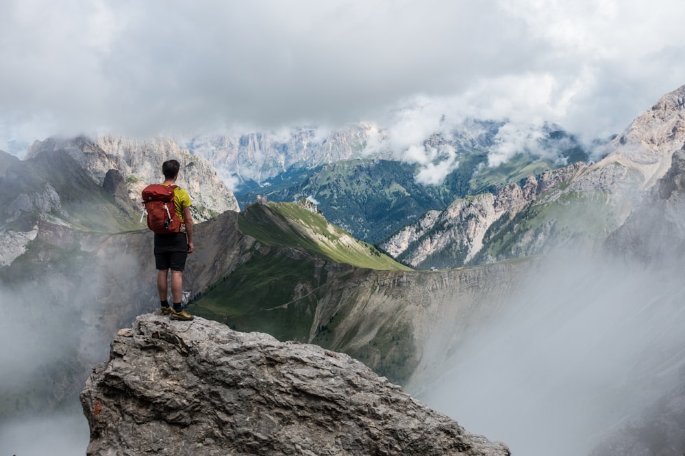 uomo con zaino rosso in piedi sulla scogliera di fronte alle montagne sotto il cielo bianco durante il giorno