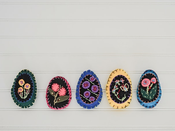 Five handmade felt egg ornaments are displayed side by side. Each egg features different embroidered floral designs, with vibrant flowers stitched onto dark backgrounds. The eggs are bordered with colorful thread, adding to their decorative appearance.