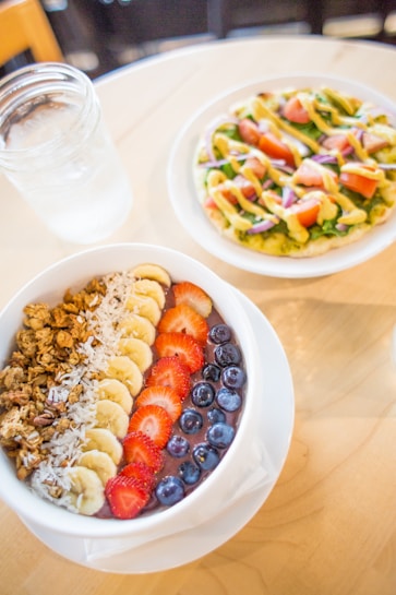 A colorful and healthy meal is placed on a light wooden table. In the foreground, there is a bowl of smoothie topped with granola, sliced bananas, strawberries, blueberries, and shredded coconut arranged in rows. In the background, a plate holds a flatbread topped with what looks like greens, sliced tomatoes, red onions, and a drizzle of dressing, possibly avocado.