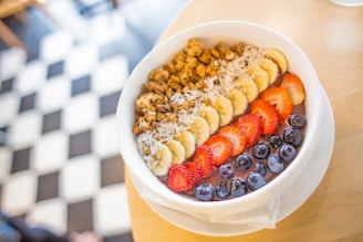 A colorful breakfast bowl topped with sliced bananas, strawberries, blueberries, granola, and shredded coconut, placed on a light wooden table with a checkered black and white floor in the background.