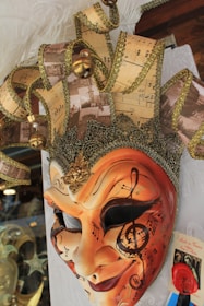 An ornate and detailed carnival mask decorated with an intricate design of musical notes and sheet music patterns. The mask is predominantly gold and orange, with glittering embellishments and a three-dimensional, textured surface. It includes a crown-like feature on top, adorned with bells, golden trim, and feathers.