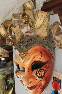 An ornate and detailed carnival mask decorated with an intricate design of musical notes and sheet music patterns. The mask is predominantly gold and orange, with glittering embellishments and a three-dimensional, textured surface. It includes a crown-like feature on top, adorned with bells, golden trim, and feathers.