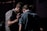 A group of people stands closely together in a dimly lit environment. One individual has their hand gently placed on another's back, suggesting a moment of comfort or support.