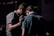 A group of people stands closely together in a dimly lit environment. One individual has their hand gently placed on another's back, suggesting a moment of comfort or support.