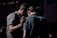 A group of people stands closely together in a dimly lit environment. One individual has their hand gently placed on another's back, suggesting a moment of comfort or support.