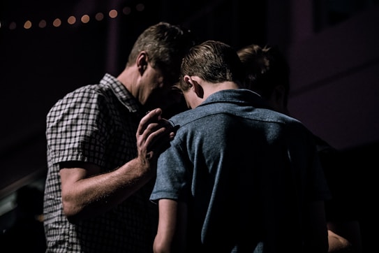 A group of people stands closely together in a dimly lit environment. One individual has their hand gently placed on another's back, suggesting a moment of comfort or support.