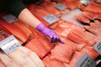 A person wearing a purple glove is pointing at a piece of fish in a display case filled with various types of raw fish fillets. Price tags with labels such as 'Red Snapper Fillet' and 'Salmon Trout' are visible.