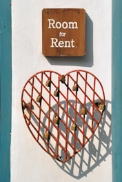 A wooden sign with white text reading 'Room for Rent' is mounted on a wall. Below the sign, there is a red metal grid in the shape of a heart, with several locks attached to its bars. The wall is painted white with a teal border.