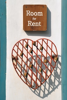 A wooden sign with white text reading 'Room for Rent' is mounted on a wall. Below the sign, there is a red metal grid in the shape of a heart, with several locks attached to its bars. The wall is painted white with a teal border.