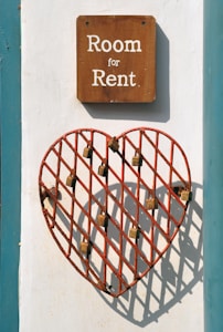 A wooden sign with white text reading 'Room for Rent' is mounted on a wall. Below the sign, there is a red metal grid in the shape of a heart, with several locks attached to its bars. The wall is painted white with a teal border.