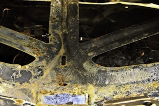 A close-up view of a rusted metallic surface, showcasing aged and corroded textures with signs of oxidation. There are circular patterns and discoloration on the metal. Loose wires and bolts are partially visible.