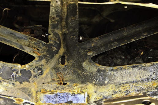 A close-up view of a rusted metallic surface, showcasing aged and corroded textures with signs of oxidation. There are circular patterns and discoloration on the metal. Loose wires and bolts are partially visible.