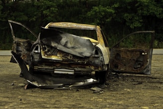 A severely burnt car is positioned in an outdoor setting with both doors open. The vehicle is heavily damaged, with charred and deformed metal surfaces. The surrounding area is a barren, dirt-covered ground with green trees in the background.