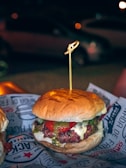 A hamburger with a juicy beef patty, melted cheese, and slices of tomato, topped with a green sauce, possibly pesto, all encased within a soft, lightly toasted bun. A wooden skewer holds it together. The burger is placed on a paper wrapper with text and graphics, creating an informal dining experience. In the background, there's a blurred, dimly lit outdoor setting with a parked car and soft bokeh lighting, suggesting nighttime.