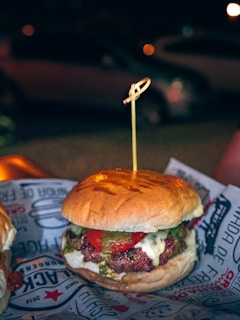 A hamburger with a juicy beef patty, melted cheese, and slices of tomato, topped with a green sauce, possibly pesto, all encased within a soft, lightly toasted bun. A wooden skewer holds it together. The burger is placed on a paper wrapper with text and graphics, creating an informal dining experience. In the background, there's a blurred, dimly lit outdoor setting with a parked car and soft bokeh lighting, suggesting nighttime.