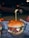 A hamburger with a juicy beef patty, melted cheese, and slices of tomato, topped with a green sauce, possibly pesto, all encased within a soft, lightly toasted bun. A wooden skewer holds it together. The burger is placed on a paper wrapper with text and graphics, creating an informal dining experience. In the background, there's a blurred, dimly lit outdoor setting with a parked car and soft bokeh lighting, suggesting nighttime.