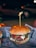 A hamburger with a juicy beef patty, melted cheese, and slices of tomato, topped with a green sauce, possibly pesto, all encased within a soft, lightly toasted bun. A wooden skewer holds it together. The burger is placed on a paper wrapper with text and graphics, creating an informal dining experience. In the background, there's a blurred, dimly lit outdoor setting with a parked car and soft bokeh lighting, suggesting nighttime.