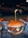 A hamburger with a juicy beef patty, melted cheese, and slices of tomato, topped with a green sauce, possibly pesto, all encased within a soft, lightly toasted bun. A wooden skewer holds it together. The burger is placed on a paper wrapper with text and graphics, creating an informal dining experience. In the background, there's a blurred, dimly lit outdoor setting with a parked car and soft bokeh lighting, suggesting nighttime.