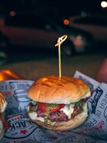 A hamburger with a juicy beef patty, melted cheese, and slices of tomato, topped with a green sauce, possibly pesto, all encased within a soft, lightly toasted bun. A wooden skewer holds it together. The burger is placed on a paper wrapper with text and graphics, creating an informal dining experience. In the background, there's a blurred, dimly lit outdoor setting with a parked car and soft bokeh lighting, suggesting nighttime.