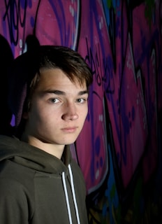 A young person with short brown hair wears a beanie and hooded sweatshirt, standing in front of a vibrant graffiti-covered wall. The graffiti features shades of pink, purple, and hints of blue and green, with abstract shapes and partial lettering.