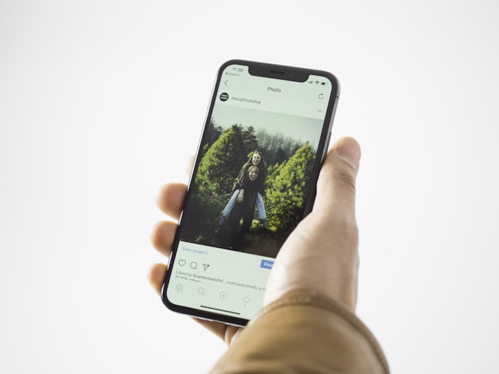A hand holding a smartphone displaying an Instagram post. The screen shows a photo of two smiling children standing in front of greenery, possibly a garden or park. The person holding the phone is wearing a brown jacket.