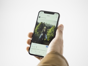 A hand holding a smartphone displaying an Instagram post. The screen shows a photo of two smiling children standing in front of greenery, possibly a garden or park. The person holding the phone is wearing a brown jacket.