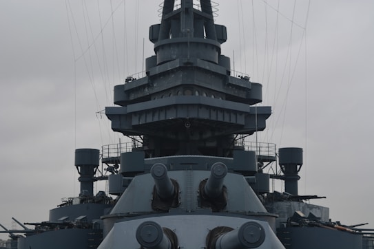 A large, formidable battleship with multiple gun turrets pointing outward. The ship has a complex superstructure with various platforms and antennas. The sky is overcast, adding a dramatic backdrop to the ship's steel-gray hull.