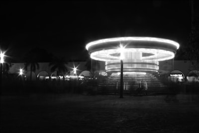 A long-exposure photograph captures a circular merry-go-round with bright lights creating rings in the darkness. In the background, there are palm trees silhouetted against a dark sky and a row of buildings with arched windows, featuring illuminated signs.