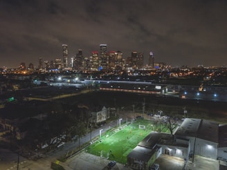 A nighttime cityscape with a brightly lit soccer field in the foreground. The field is surrounded by industrial buildings and a neighborhood, while the city skyline with tall, illuminated skyscrapers is visible in the background. The clouded sky adds to the urban ambiance.