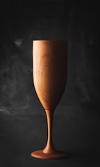 A wooden goblet stands against a dark background. The goblet's finish showcases the natural grain and texture of the wood, highlighting its handcrafted appearance.