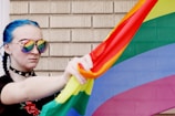 A person with blue braided hair wears sunglasses reflecting a rainbow flag. They hold the colorful flag against a brick wall backdrop. The person wears a choker with star designs and a black top with floral embroidery.