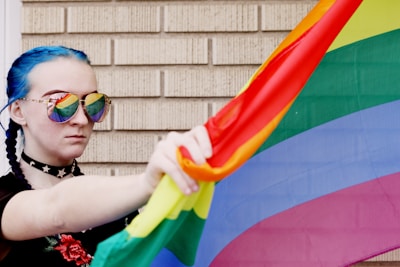 A person with blue braided hair wears sunglasses reflecting a rainbow flag. They hold the colorful flag against a brick wall backdrop. The person wears a choker with star designs and a black top with floral embroidery.