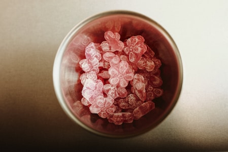 A glass container filled with pink, flower-shaped candies viewed from above. The candies have a powdery coating, and the surface they are on is a neutral tone.