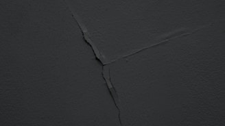 A dark, cracked wall with a visible diagonal fracture running across the surface. The texture is rough and the lighting is dim, giving it a shadowy appearance.