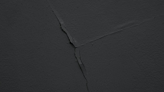 A dark, cracked wall with a visible diagonal fracture running across the surface. The texture is rough and the lighting is dim, giving it a shadowy appearance.
