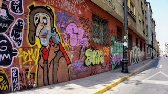 A street scene features a long wall covered in vibrant graffiti art, including a large cartoonish face with a bird perched on its shoulder, surrounded by various colorful tags and text. The building's wall is red with barred windows, and a black street lamp stands on the sidewalk. The street is lined with buildings and continues into the distance.