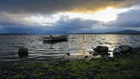 A serene lakeside scene featuring a small, weathered boat floating on the water. The sky is filled with dramatic clouds, illuminated by the setting or rising sun, casting a warm glow on the landscape. The foreground includes a rocky shore with patches of green moss, and distant hills are visible across the horizon.