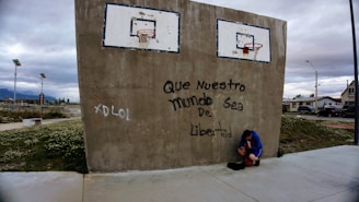 A concrete wall features two basketball hoops with faded paint, and graffiti reads 'Que Nuestro Mundo Sea De Libertad.' A person in a blue jacket and shorts is seated on the ground, holding a basketball. This scene takes place in an urban setting with cloudy skies and residential houses in the background.