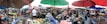 A bustling outdoor marketplace with numerous vendors and shoppers under colorful umbrellas. Various goods like fruits, vegetables, and other items are displayed on tables. People are interacting and engaging in commerce, with a lively atmosphere and a sense of community.