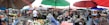 A bustling outdoor marketplace with numerous vendors and shoppers under colorful umbrellas. Various goods like fruits, vegetables, and other items are displayed on tables. People are interacting and engaging in commerce, with a lively atmosphere and a sense of community.