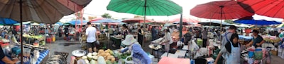 A bustling outdoor marketplace with numerous vendors and shoppers under colorful umbrellas. Various goods like fruits, vegetables, and other items are displayed on tables. People are interacting and engaging in commerce, with a lively atmosphere and a sense of community.