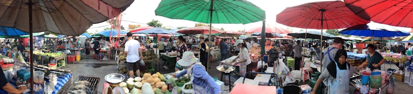 A bustling outdoor marketplace with numerous vendors and shoppers under colorful umbrellas. Various goods like fruits, vegetables, and other items are displayed on tables. People are interacting and engaging in commerce, with a lively atmosphere and a sense of community.