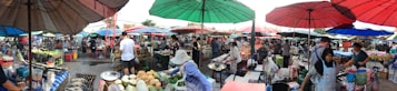 A bustling outdoor marketplace with numerous vendors and shoppers under colorful umbrellas. Various goods like fruits, vegetables, and other items are displayed on tables. People are interacting and engaging in commerce, with a lively atmosphere and a sense of community.