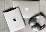A silver laptop and a tablet, both featuring Apple logos, lie on a textured gray surface. Nearby, a pair of silver over-ear Bose headphones rests beside them.