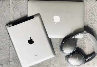 A silver laptop and a tablet, both featuring Apple logos, lie on a textured gray surface. Nearby, a pair of silver over-ear Bose headphones rests beside them.