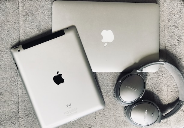 A silver laptop and a tablet, both featuring Apple logos, lie on a textured gray surface. Nearby, a pair of silver over-ear Bose headphones rests beside them.